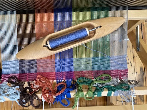 A wooden loom with a blue thread spindle in the center, set against a colorful woven textile with vertical stripes in green, blue, red, orange, and white.