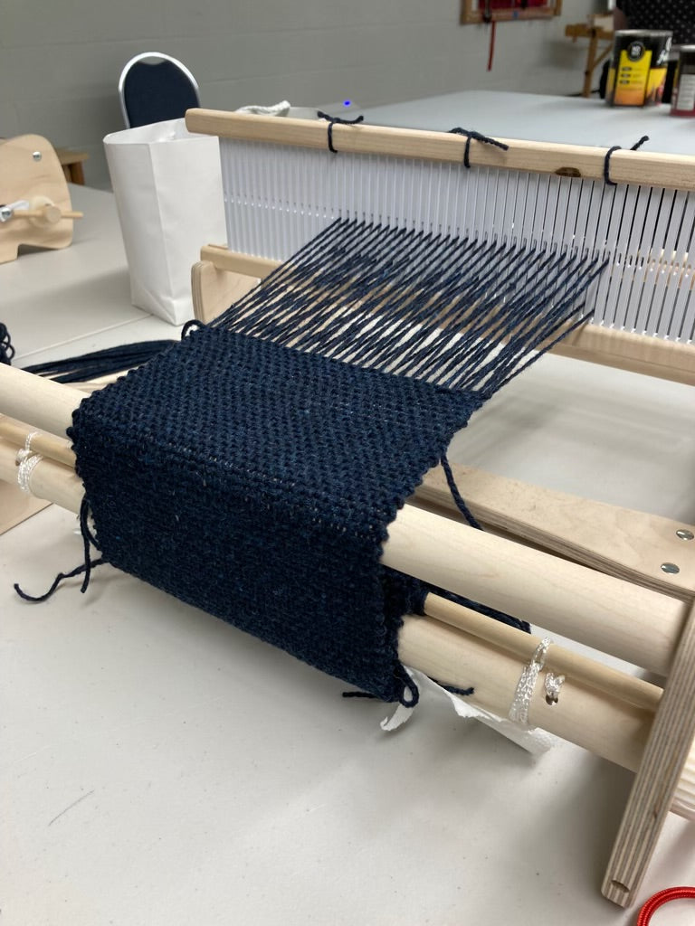 A close-up of a dark navy blue knitted fabric draped over a wooden loom, with warp threads visible and part of a textile workshop or studio in the background.
