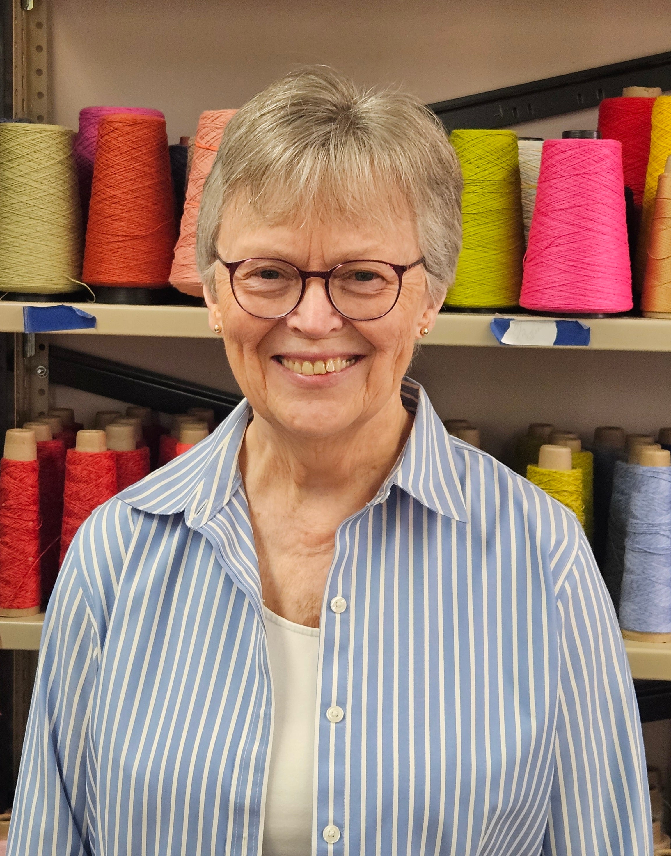 A smiling woman with short gray hair and glasses, wearing a light blue and white striped button-up shirt over a white top, standing in front of shelves filled with colorful spools of thread.