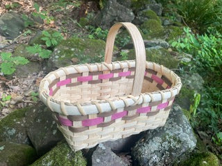 A handwoven basket with a natural beige wood tone and pink woven accents, featuring a single curved handle, set outdoors on mossy rocks with green foliage in the background.