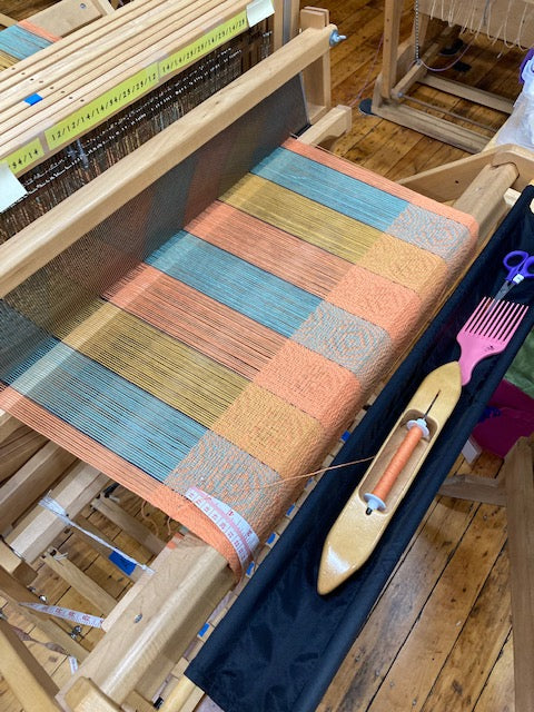 A wooden loom with a colorful woven textile featuring vertical stripes in peach, teal, beige, and brown. A pink comb tool and wooden spool are resting on the loom, set against a rustic wooden floor background.