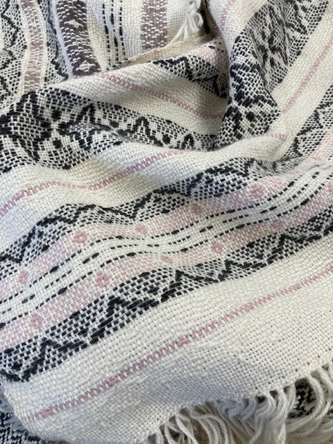 A close-up of a woven textile with fringe, featuring a cream base and horizontal bands of black geometric patterns and light pink dotted lines.