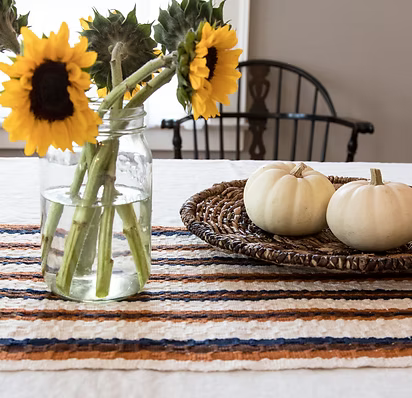 A rustic woven table runner in orange, beige, and navy blue stripes is styled on a white tablecloth. On the left, a clear glass mason jar holds sunflowers and thistle stems, while on the right, a round woven tray holds two small cream pumpkin