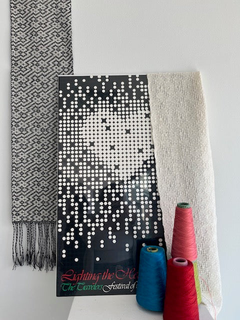 A woven textile sample with a black-and-white pixelated pattern and gray-and-white herringbone fringe, displayed on a white background with three colored spools of thread above it.